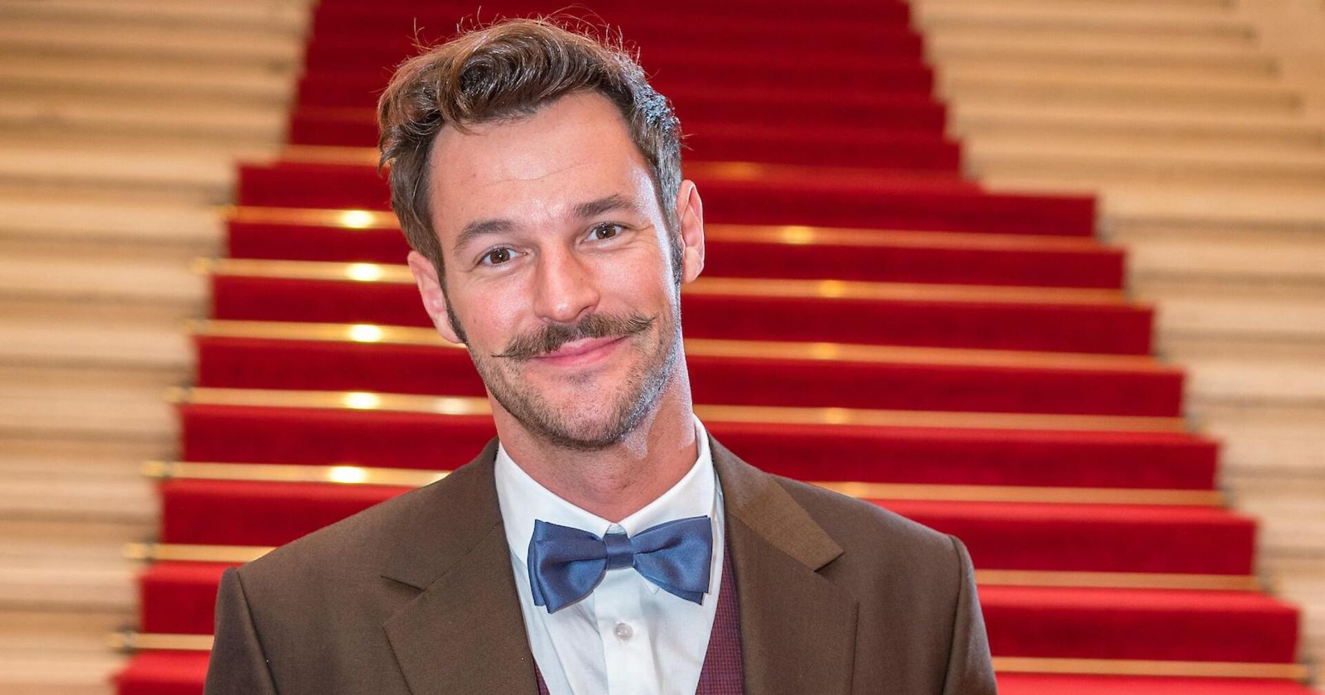 Paul Pizzera mit einem braunen Smoking vor einem Treppenaufgang.