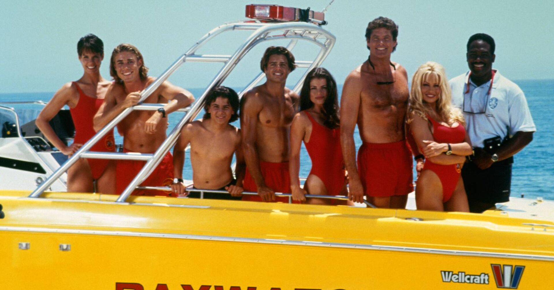 Das Baywatch-Ensemble, das in einer Reihe auf einem gelben Motorboot posiert