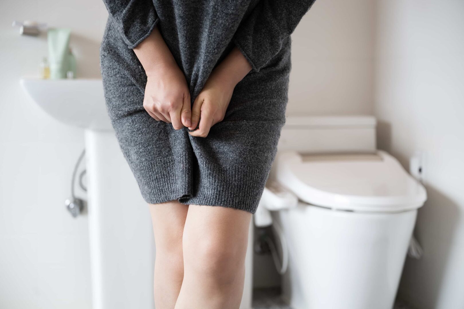 Eine Frau muss dringend auf die Toilette. Sie zwickt die Beine zusammen. Im Hintergrund die rettende WC-Schüssel.
