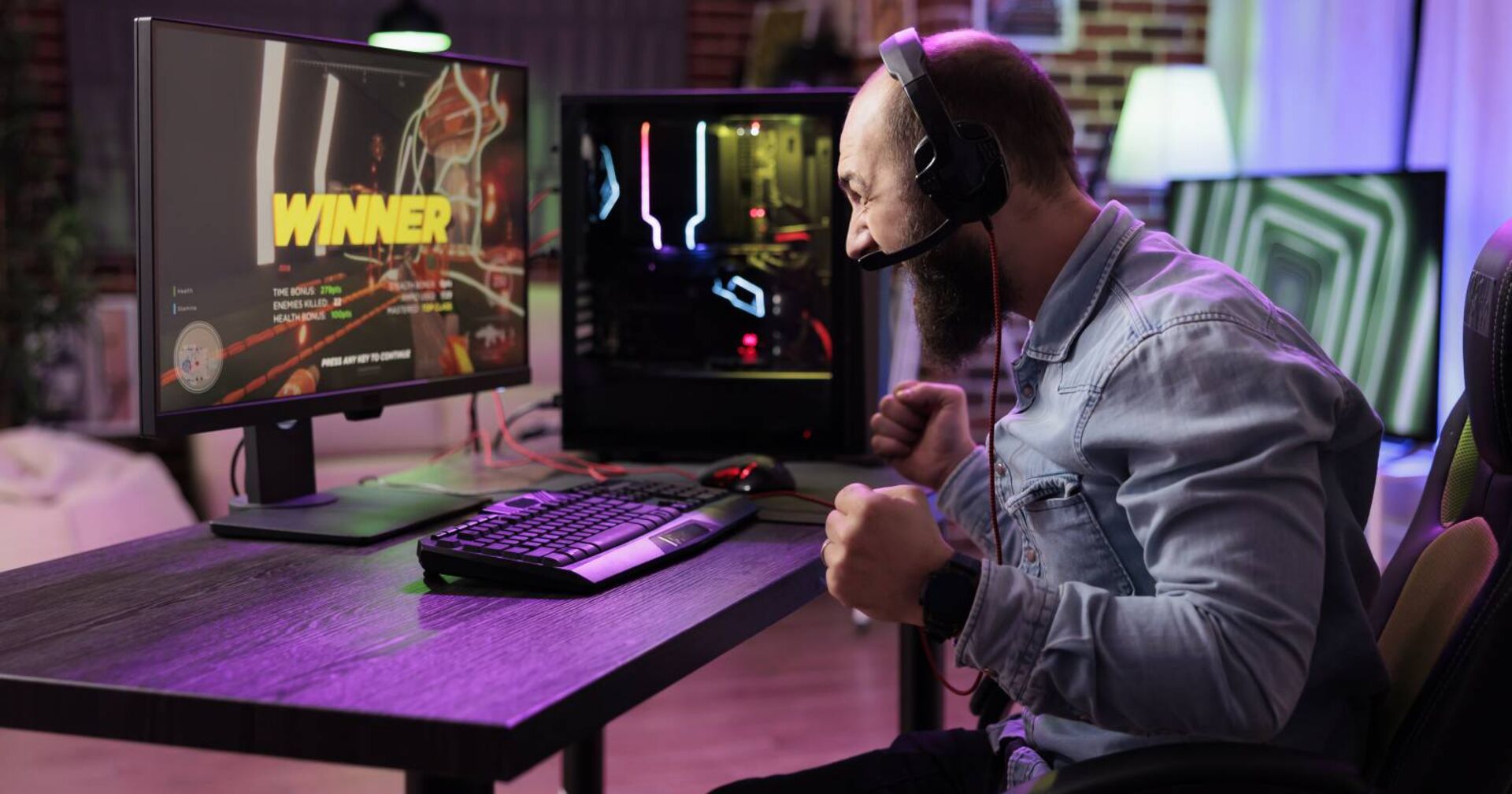 Mann, der mit Kopfhörern vor dem PC sitzt und ein Spiel gewonnen hat. Auf dem Monitor des PCs ist in bunten Buchstaben WINNER, zu deutsch Sieger, zu lesen.