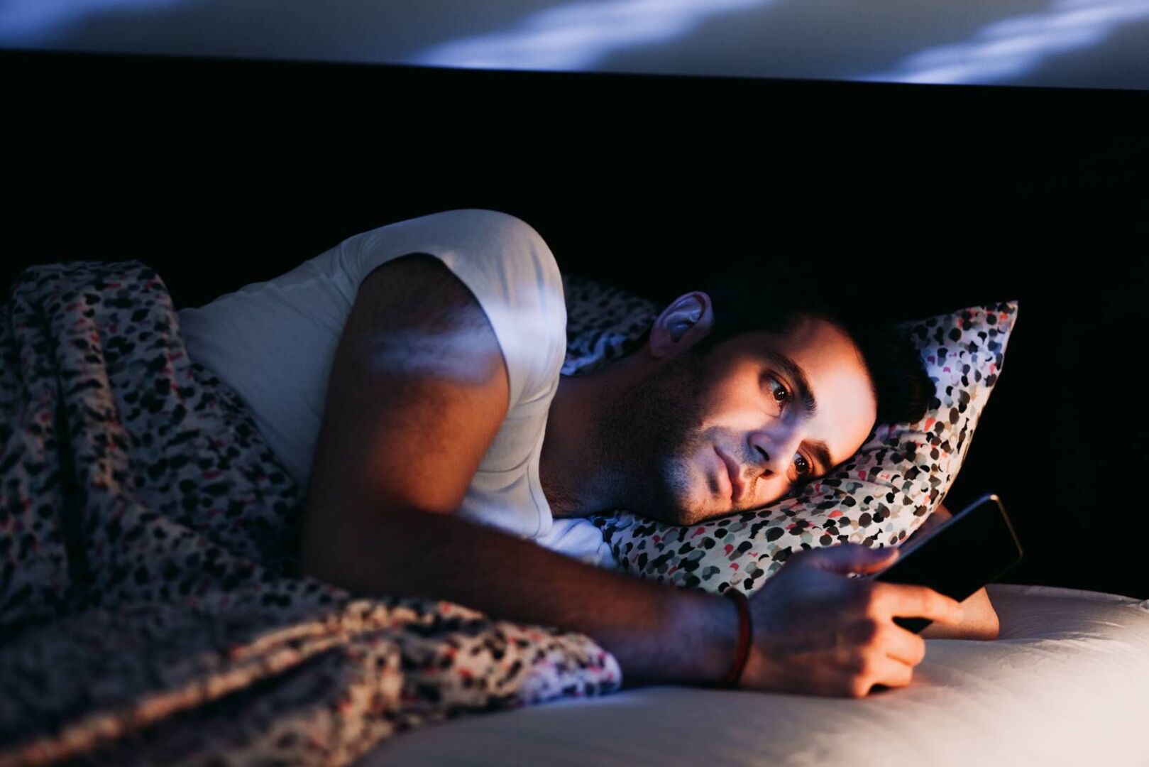 Junger Mann liegt auf der Seite im Bett und schaut auf das Display seines Handys.