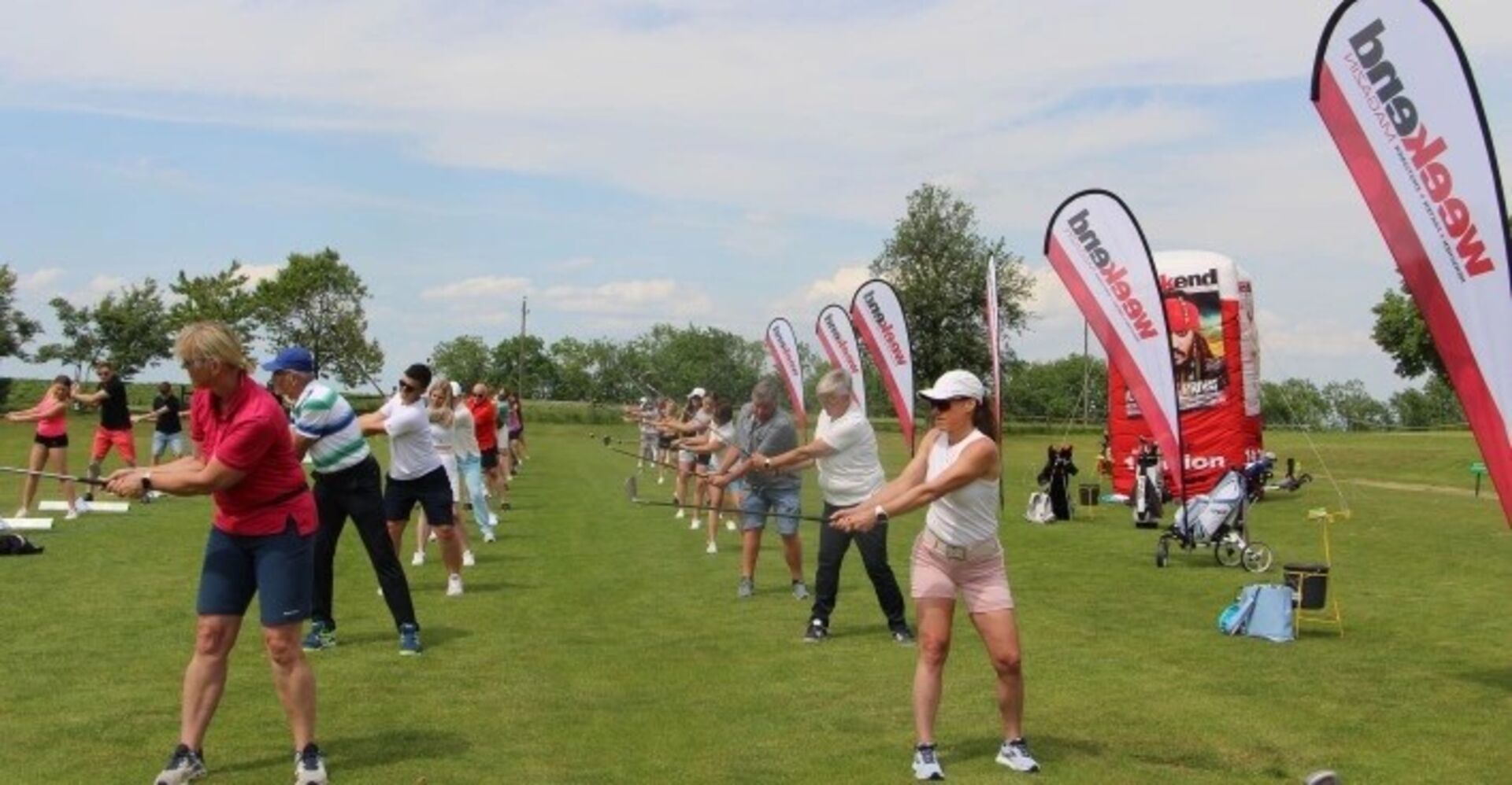 Eine Gruppe von Golfanfängern versucht sich beim Abschlag. Rechts davon sind Fahnen mit der Aufschrift www.weekend.at zu sehen