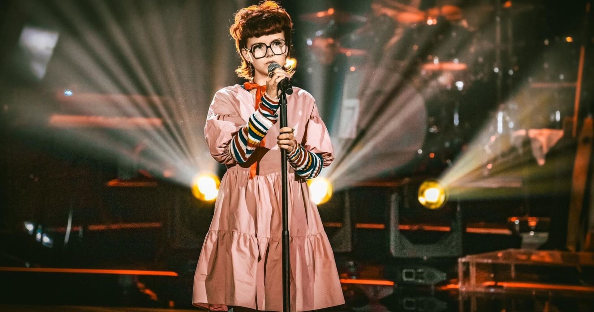 Victoria Knogler auf der steht auf der Bühne von "The voice kids." Die Sängerin trägt ein rosa Kleid und einen Pullover mit bunten Querstreifen. Ihre braunen Haare sind kurz geschnitten, ihre Augen sieht man durch eine viereckige Brille. In der Hand hält sie ein Mikrofon.