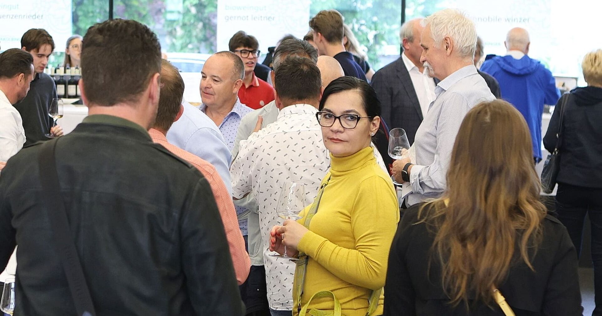 Eine Gruppe von Menschen beim Weinwunder in Linz. In der Bildmitte: Eine Dame mit schwarzen Haaren, gelbem Pullover und Brille hält ein Glas Wein.