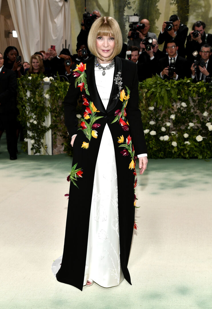 Eine Frau mit Bob in einem weißen Kleid, darüber trägt sie einen schwarzen Mantel mit Blumen