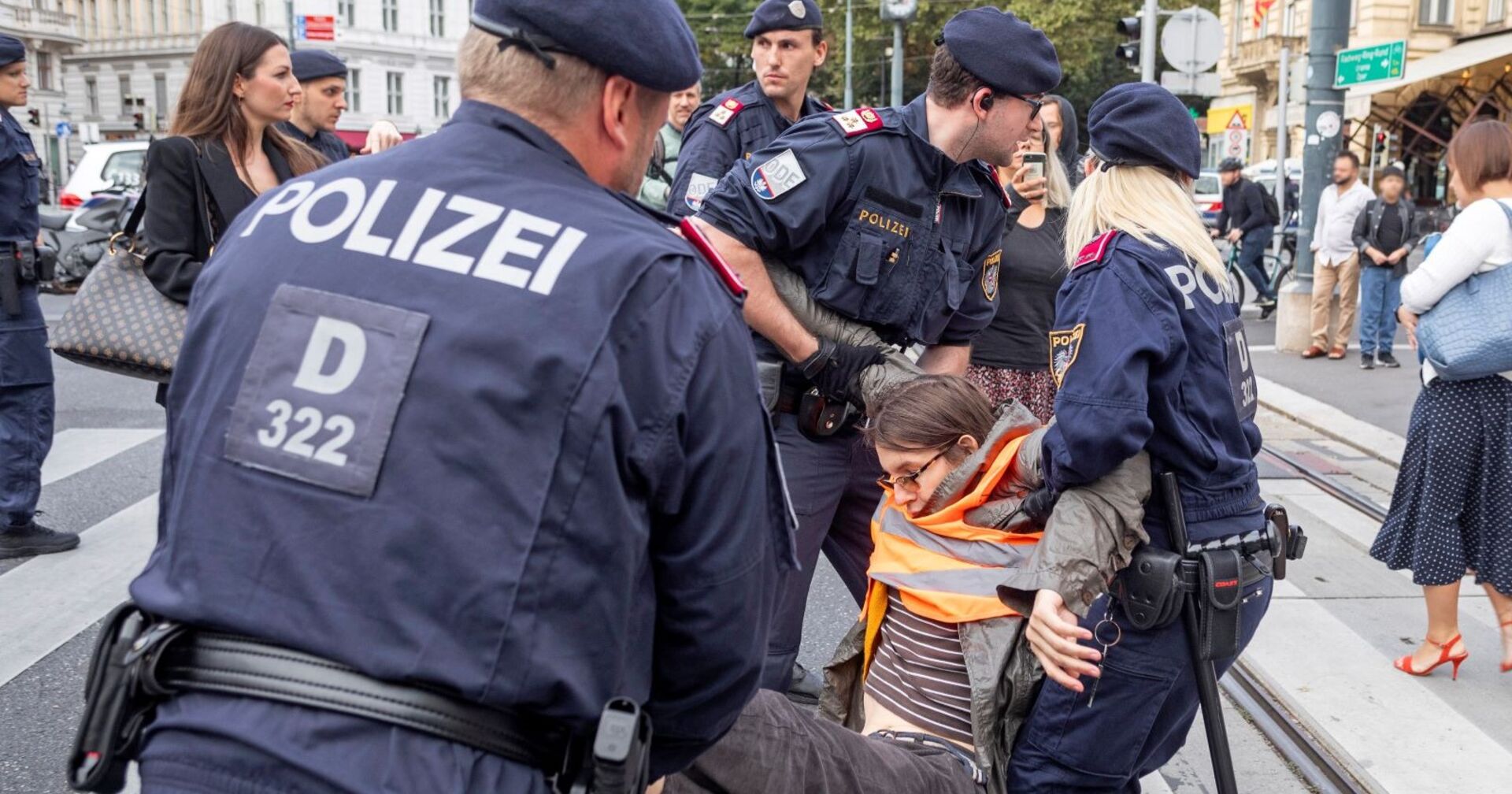 Drei Polizisten tragen eine Aktivistin in einer orangen Warnweste weg.