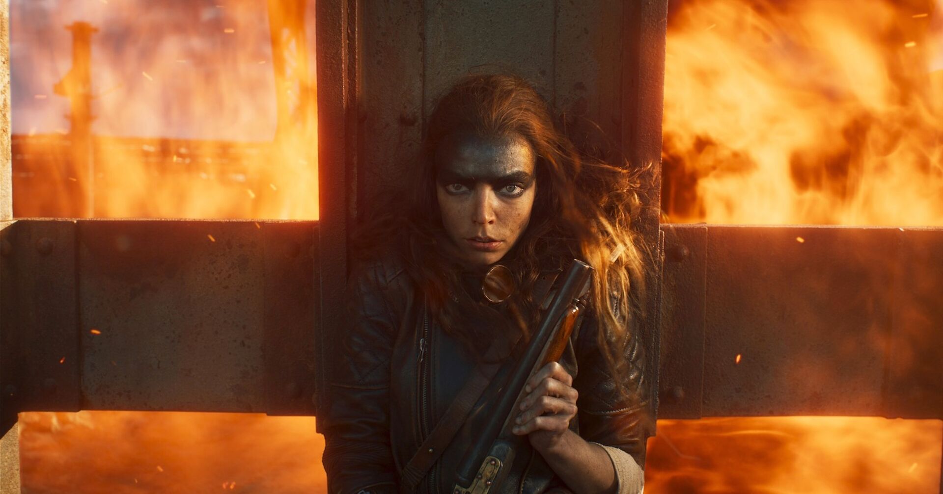 Eine Schauspielerin vom Film "Furiosa: A Mad Max Saga" mit viel schwarzer Schimnke. Hinter ihr sind Flammen. Sie trägt eine Waffe.