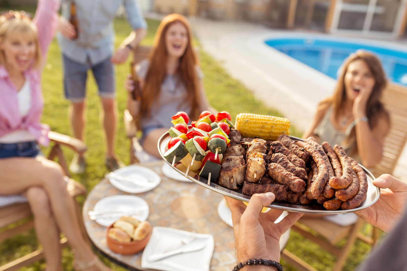 Drei lachende Frauen freuen sich auf das Essen: Auf einer Platte liegen gegrillte Gemüsespieße, Maiskolben, Cevapcici  und Würstel.