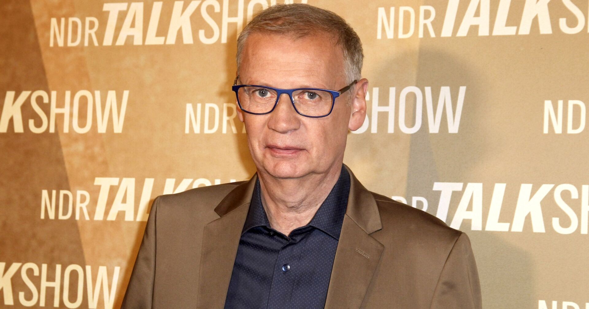 Günther Jauch in mit einem beigen Sakko vor einer beigen Fotowand.
