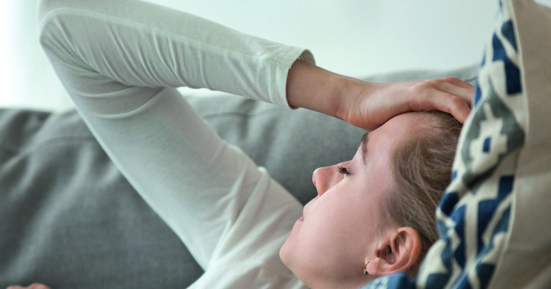 Eine junge Frau mit blonden Haaren liegt auf einer grauen Couch. Sie trägt eine blaue Jean und einen weißen Pullover. Ihr Kopf ruht auf einem Polster. Eine ihre Hände ruht auf der Stirn, da der Kopf schmerzt.