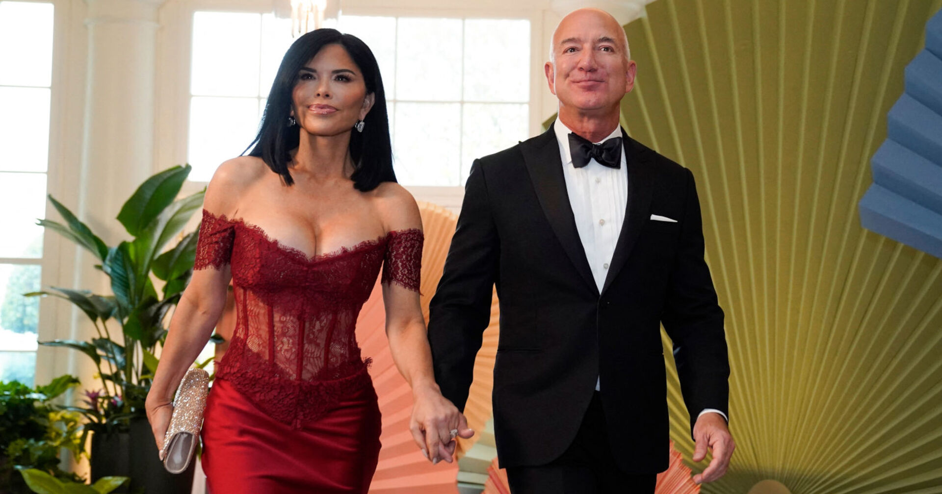 Eine Frau in einem tief ausgeschnittenen roten Kleid hält Händchen mit einem Mann in einem Smoking