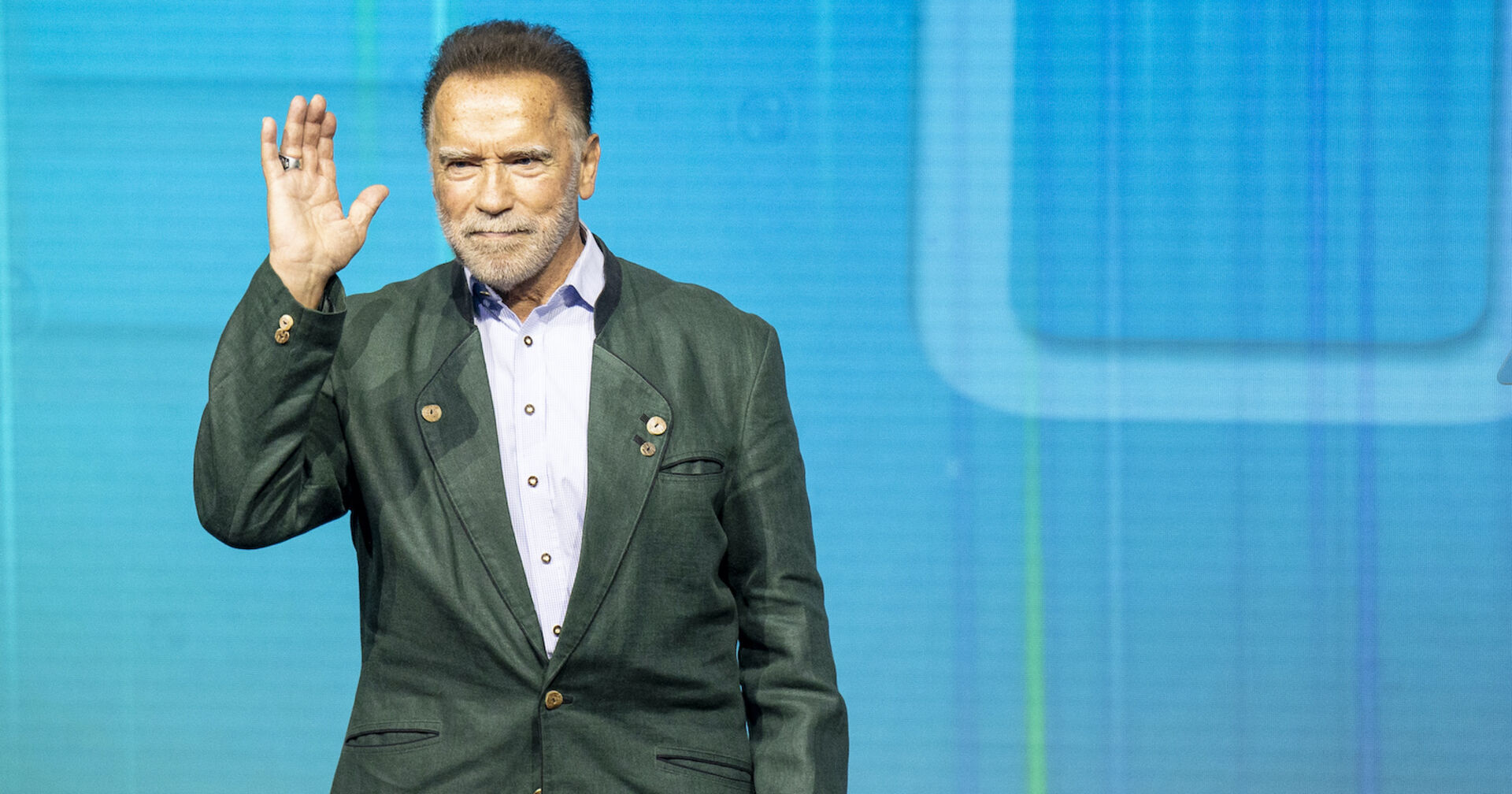 Arnold Schwarzenegger winkt in einem grünen Sakko von der Bühne.