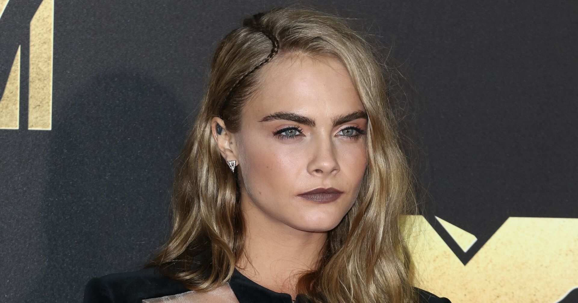 Cara Delevigne mit offenen blonden Haaren und einem dunklen Lippenstift vor einer Fotowand.
