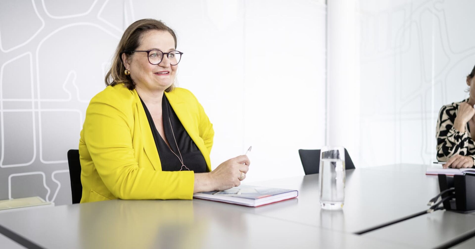 Anette Klinger, Miteigentümerin von Internorm sitzt an einem Schreibtisch auf der ein Notizblock liegt und ein Glas mit Wasser steht. Sie trägt eine braune Brille ein gelbes Sakko und eine schwarze Bluse.