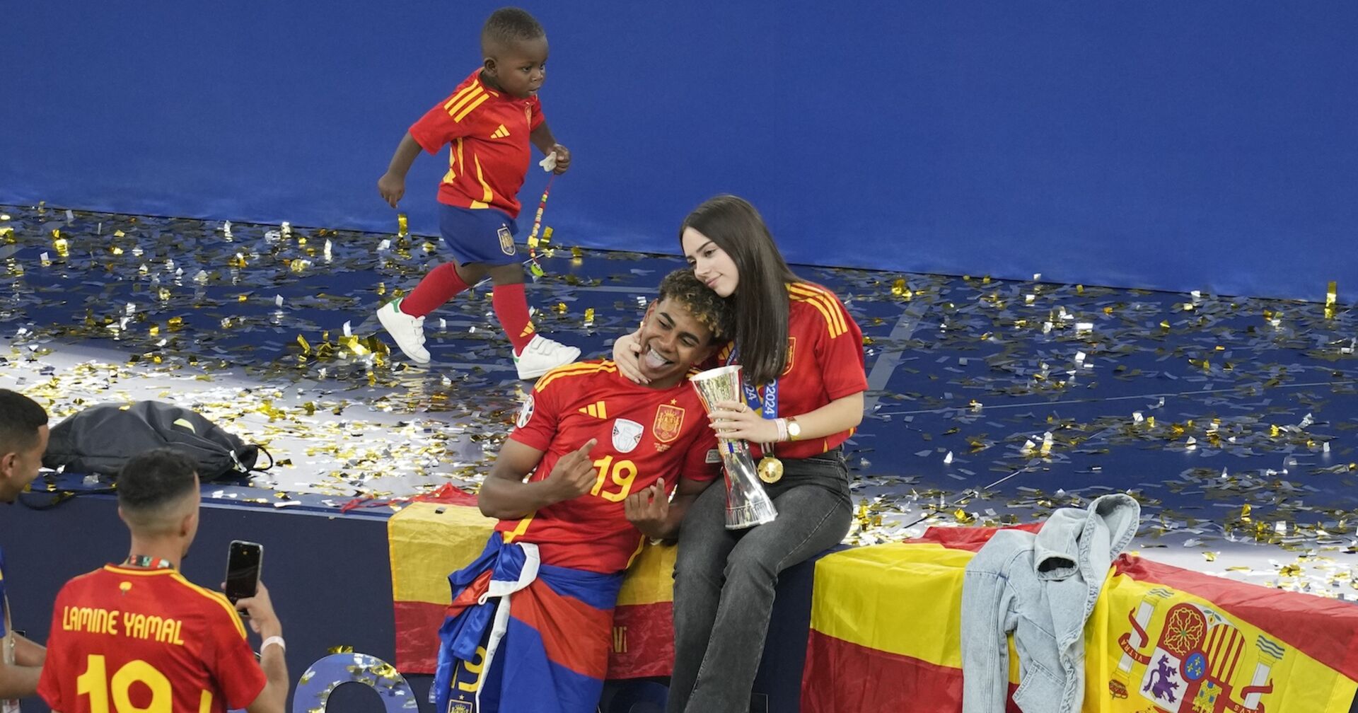 Yamal posiert mit seiner Freundin für ein Selfie. Sie hat zärtlich den Arm um seinen Nacken gelegt und trägt seine Medaille und Pokal. Im Hintergrund läuft ein Kleinkind - Yamals Bruder - durchs Bild.