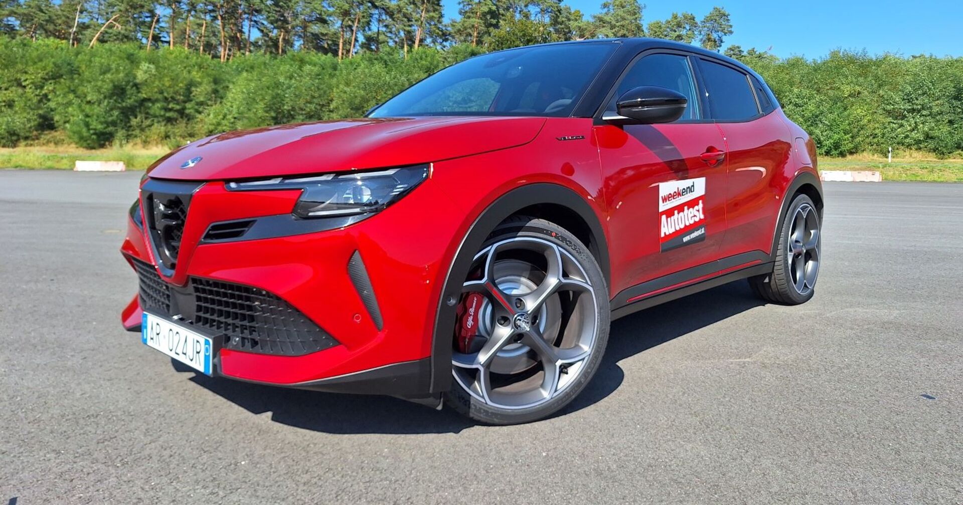 Ein roter Alfa Junior steht auf einem asphaltierten Parkplatz. das Auto glänzt in der Sonne, im Hintergrund sind Föhren und andere Bäume zu erkennen.