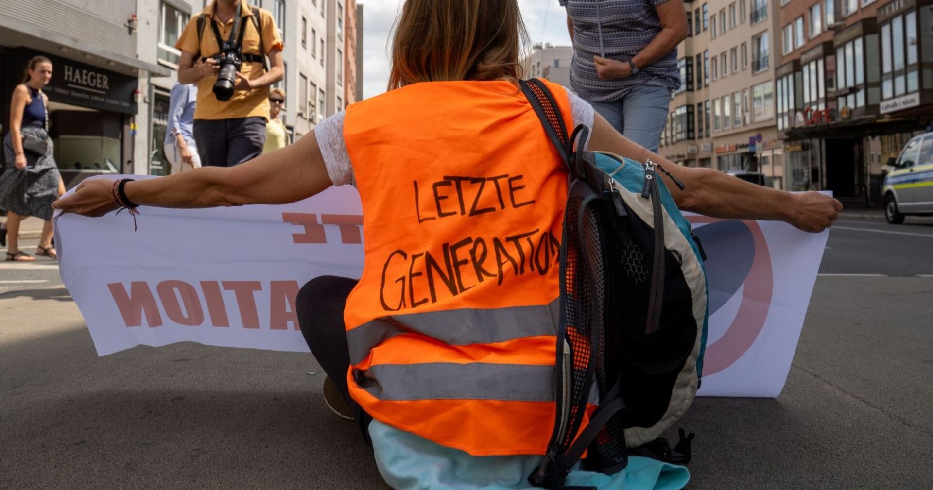 Eine Teilnehmerin der Protestbewegung Letzte Generation mit einer Warnweste und einem Transparent auf der Straße sitzend.