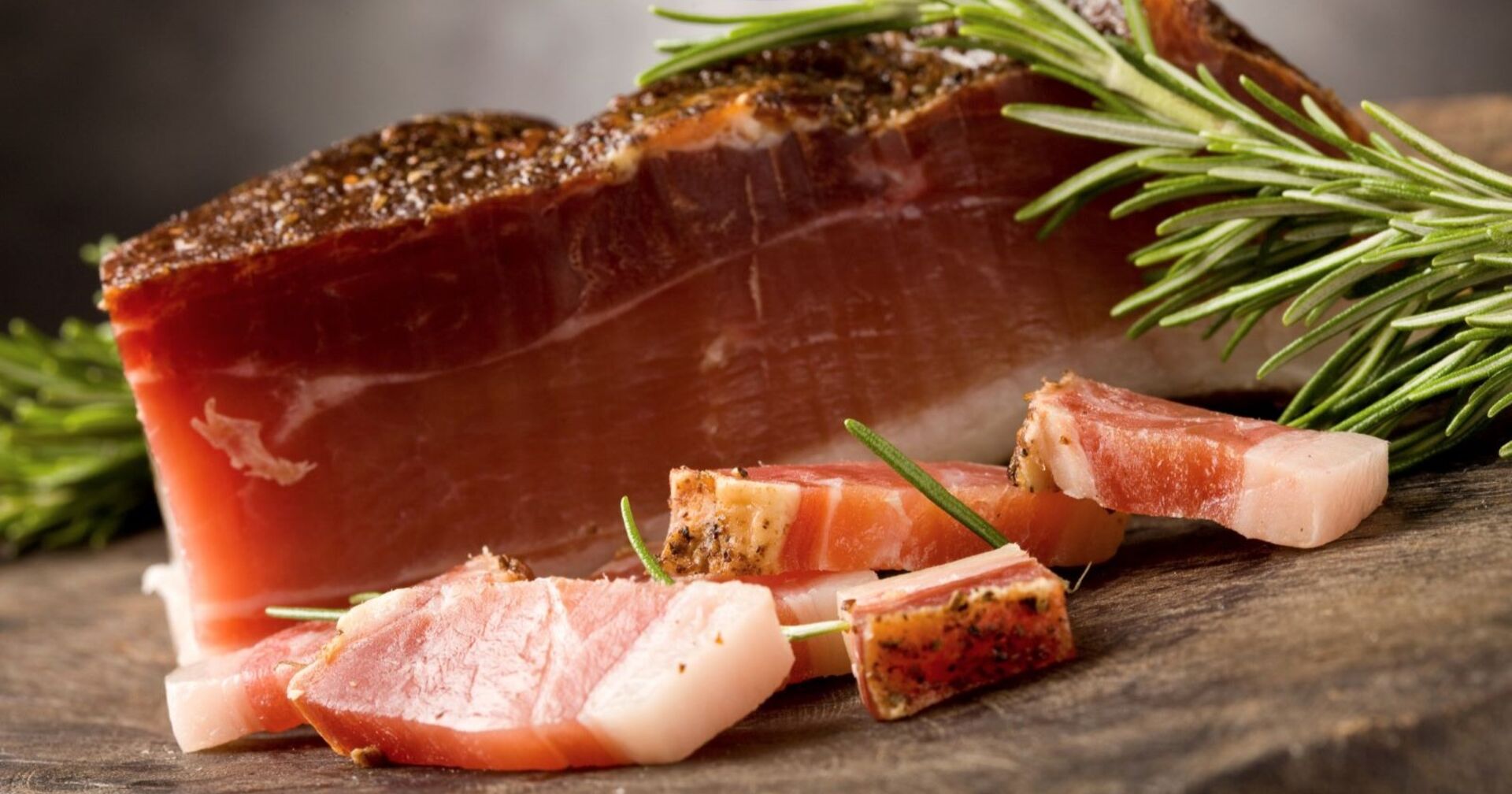 Ein Stück magerer Speck liegt auf einem Holzbrett. Davor liegen einige dünne Streifen Speckt. Zu sehen ist auch ein Rosmarinzweig, der den Speck verschönert.