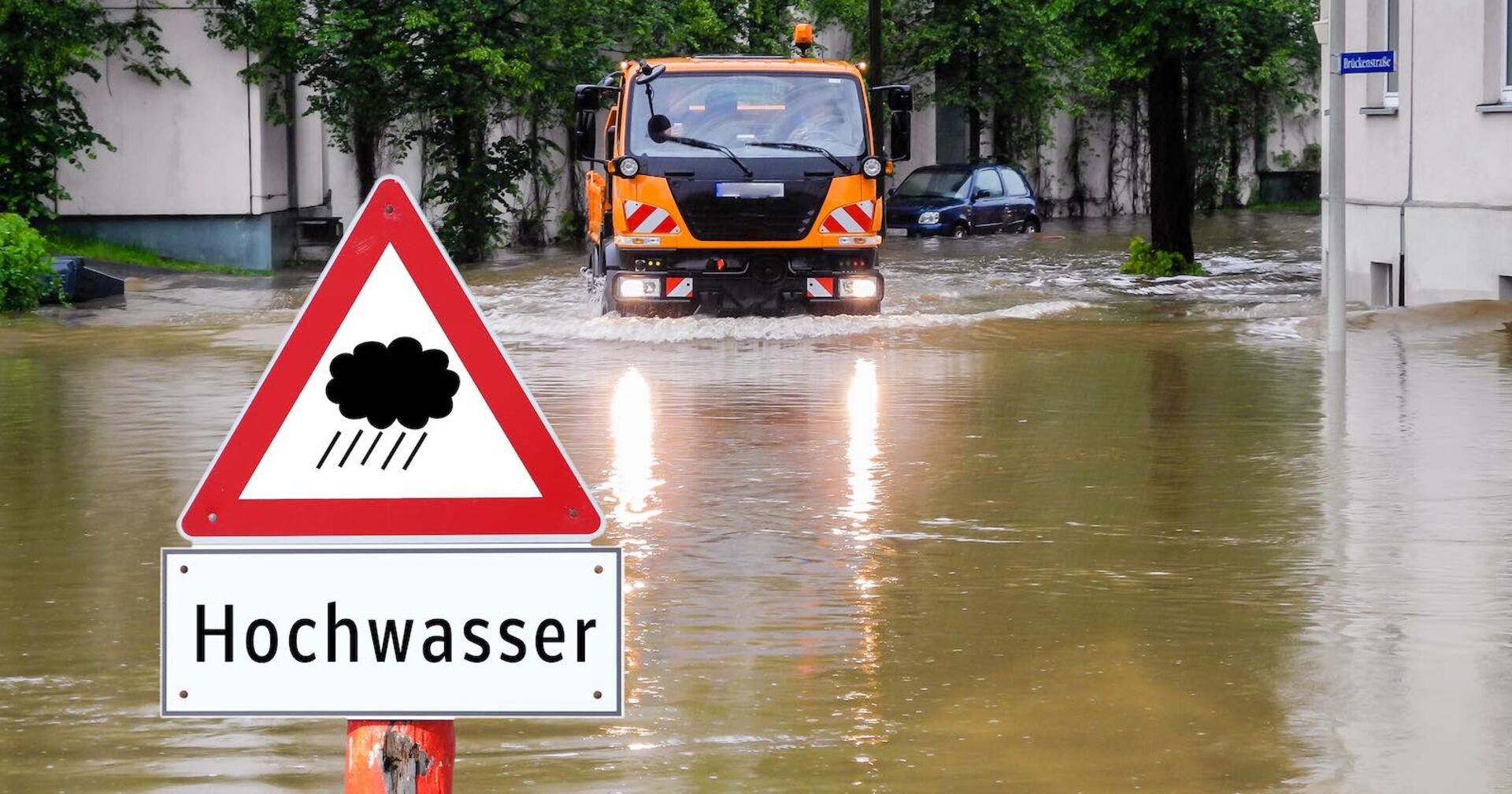 Hochwasser: Gemeinde-Lastwagen auf überschwemmter Straße und ein Hinweisschild "Hochwasser".