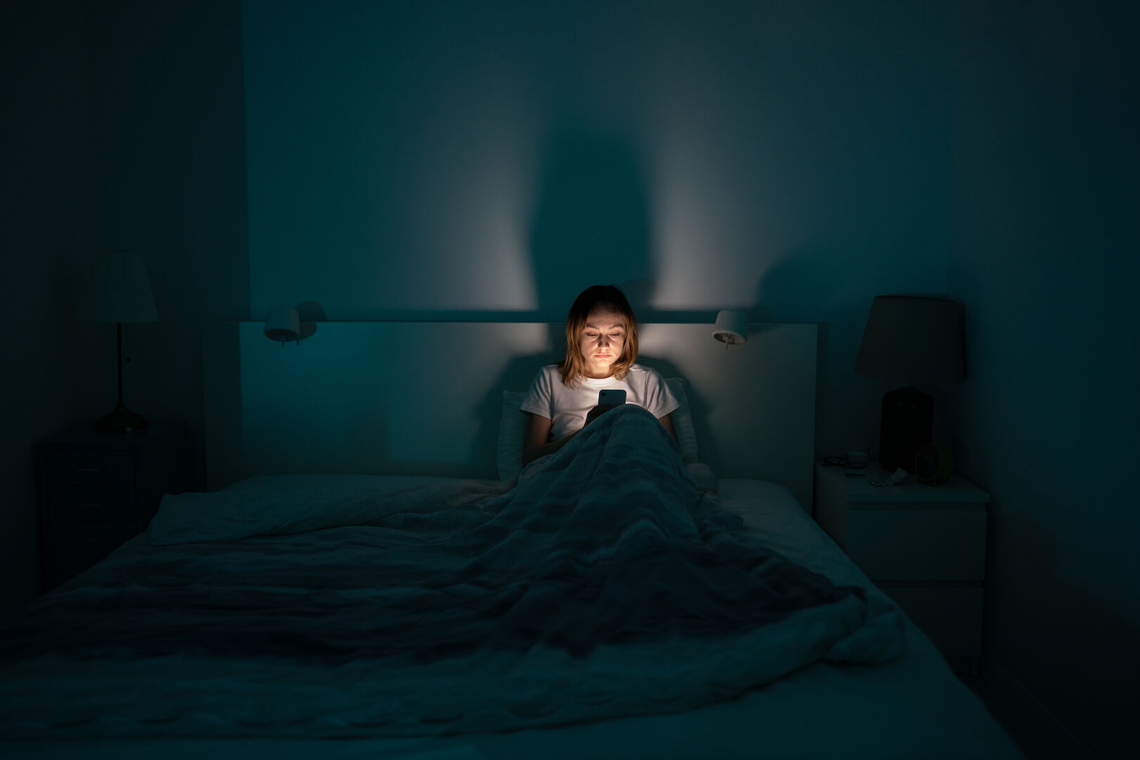 Eine Frau, die in einem dunklen Schlafzimmer in ihrem Bett sitzt und auf ihr Handy, dessen Bildschirmlicht ihr Gesicht erhellt, starrt