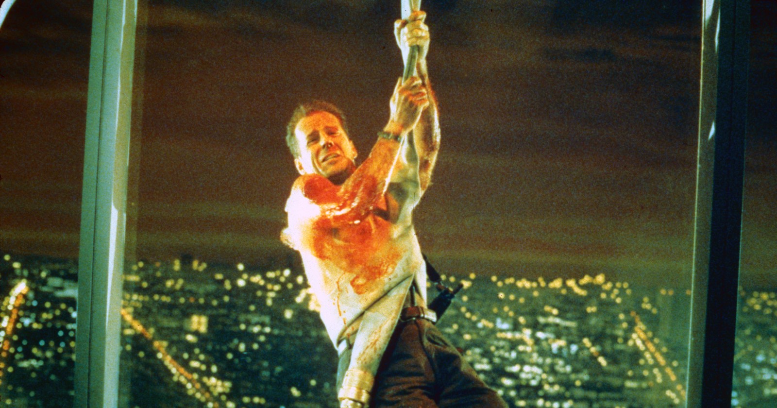 Eine Szene aus "Stirb langsam", in der der blutende Bruce Willis an einem Feuerwehrschlauch vor der Fensterfront eines Hochhauses baumelt
