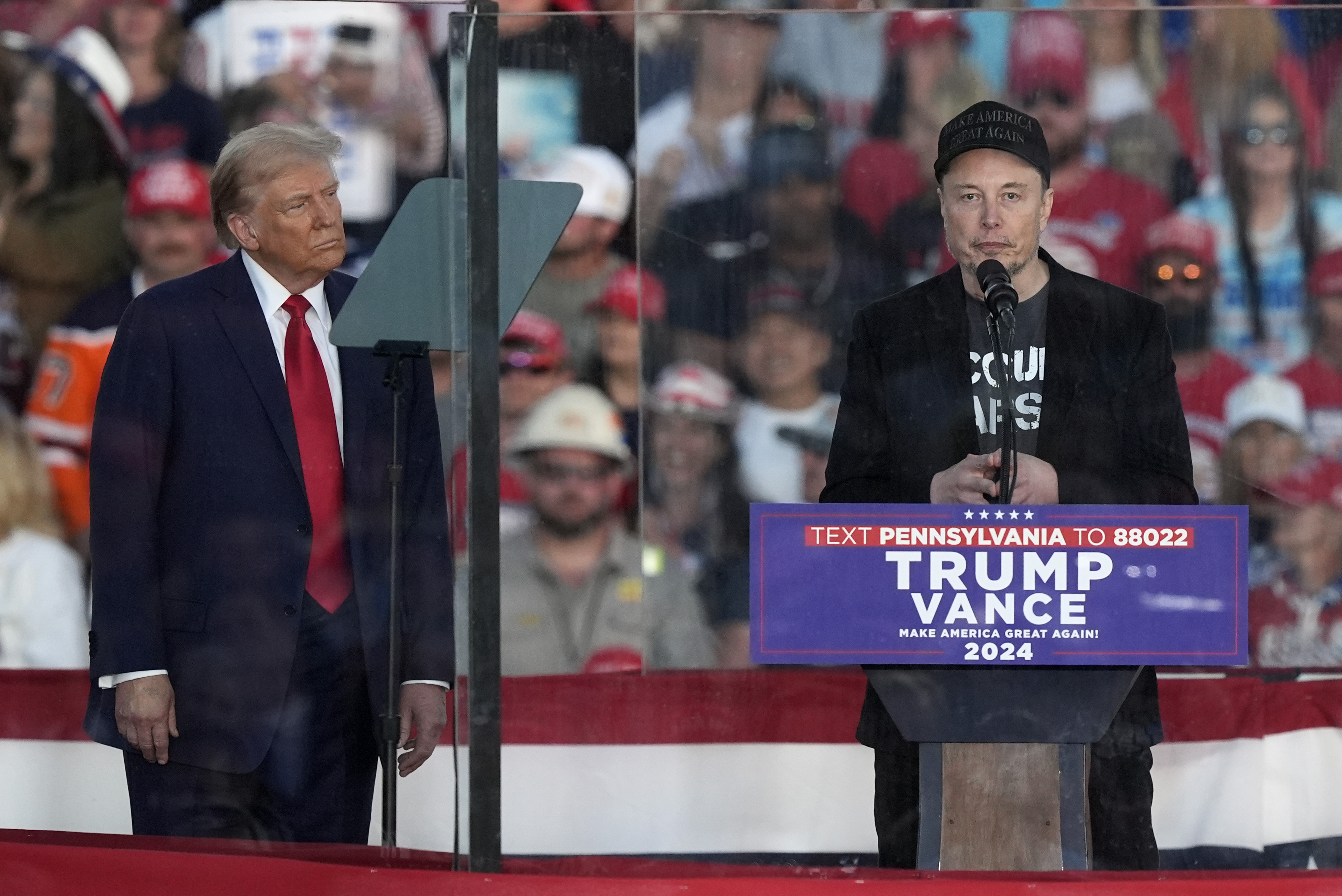 Elon Musk, der hinter einem Rednerpult auf einer Wahlkampfveranstaltung Donald Trumps steht