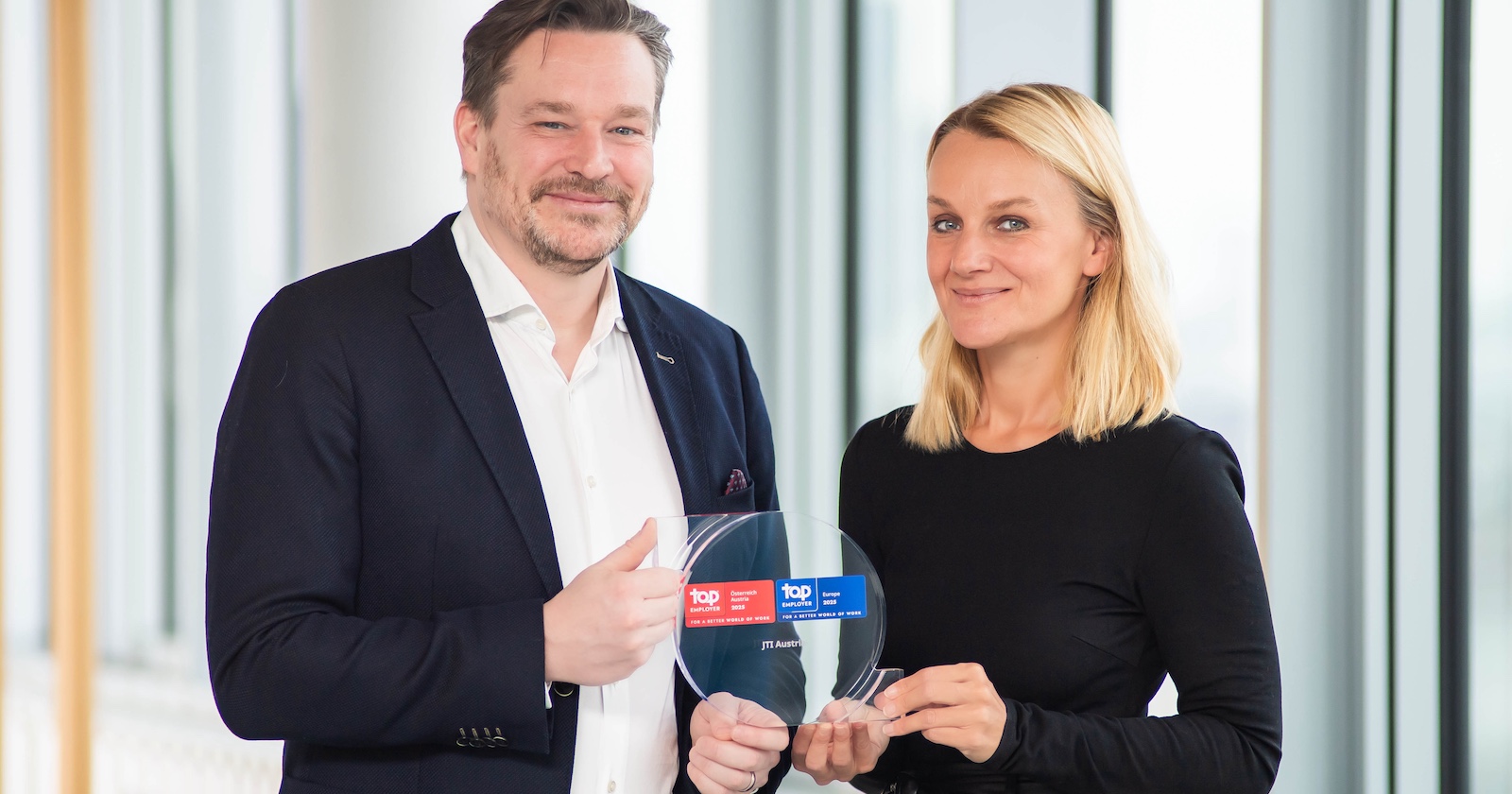 Ein Mitarbeiter und eine Mitarbeiterin von JTI Austria präsentieren eine Glastrophäe