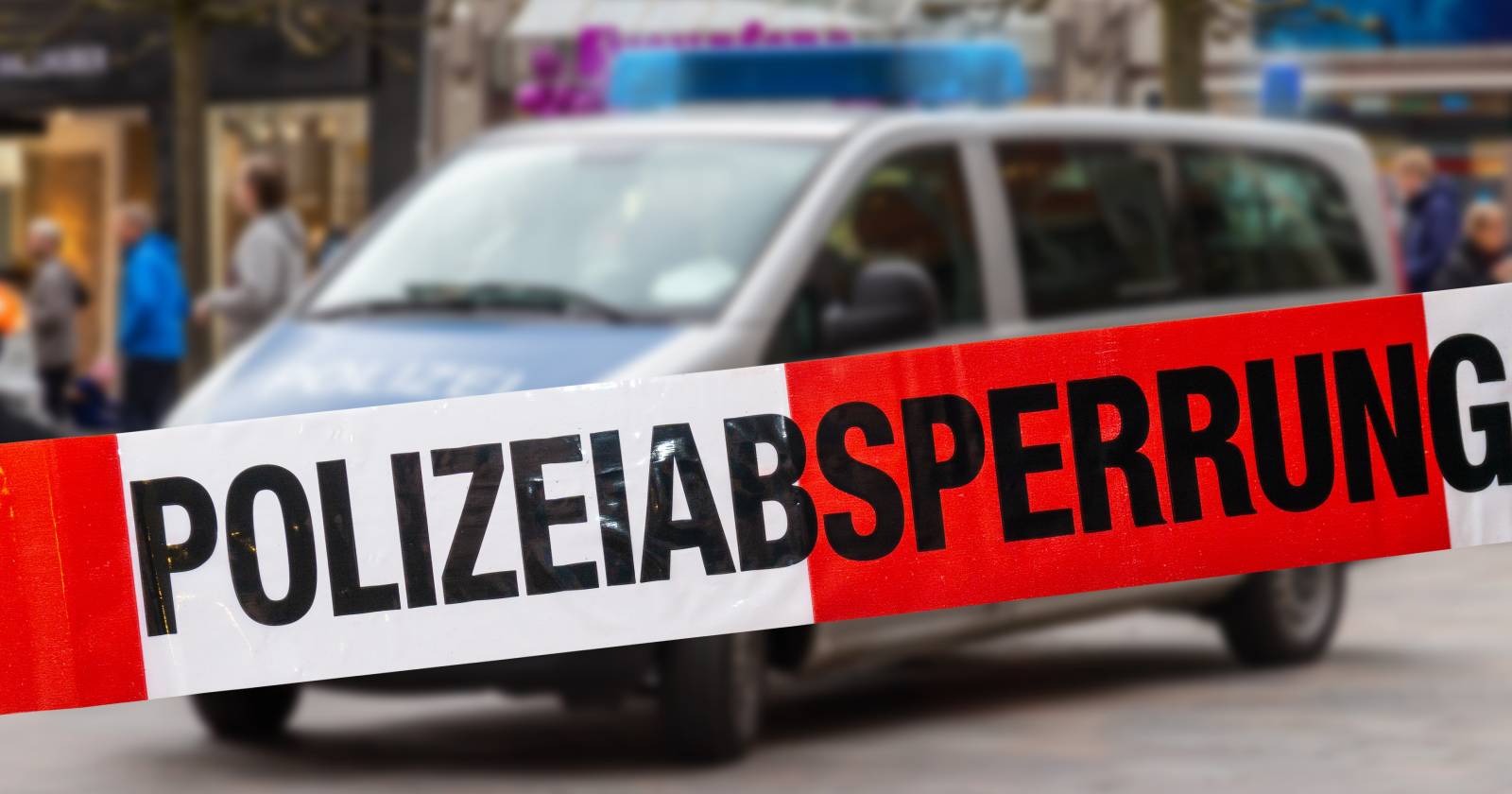 Ein Absperrband auf dem der Schriftzug "Polizeiabsperrung" zu lesen ist und in dessen Hintergrund sich ein leicht verschwommen erscheinendes Polizeiauto befindet