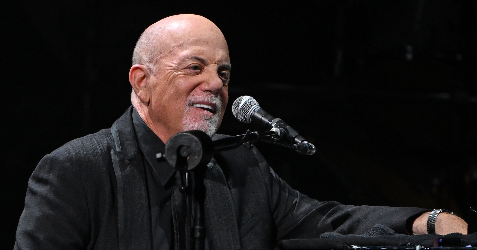 Billy Joel sitzt im dunklen Bühnenlicht an einem Mikrofon und lächelt. Er trägt ein schwarzes Sakko und eine Uhr am Handgelenk.