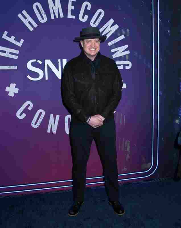 Brendan Fraser ganz in schwarz gekleidet mit Hut, lächelt in die Kamera, die Hände hat er vor seinem Körper zusammengeschlossen.