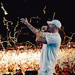 Der Hip-Hop-Musiker RAF Camora performt auf einer Bühne bei Nacht, mit Mikrofon in der Hand und vor einem jubelnden Publikum. Effekte fliegen durch die Luft.