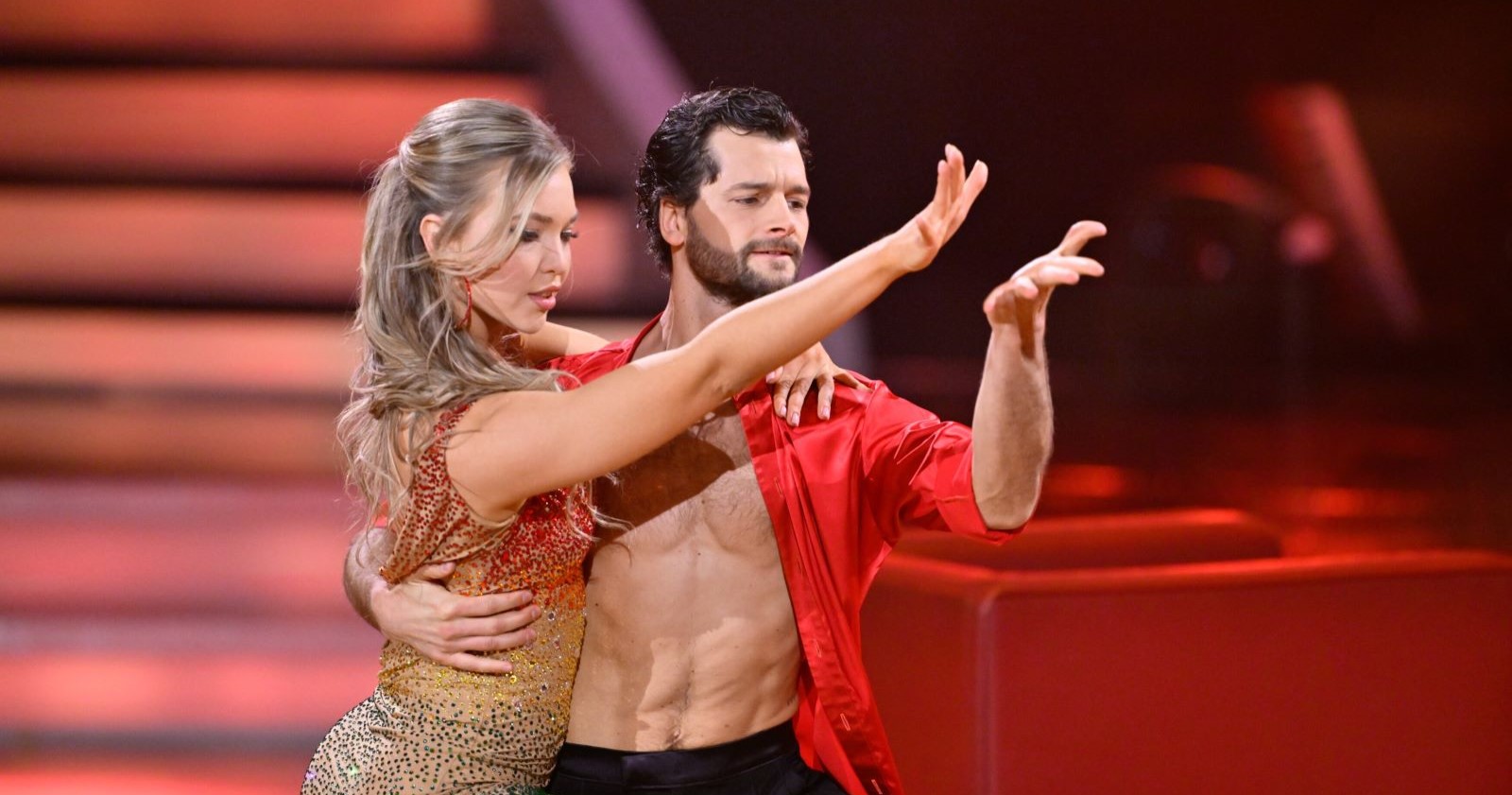 Aaron Karl tanzt barbrüstig in rotem Hemd mit seiner Tanzpartnerin Katya Mizera im Glitzerkleid. Beide zeigen eine ausdrucksstarke Pose bei der Rumba auf der „Dancing Stars“-Bühne.