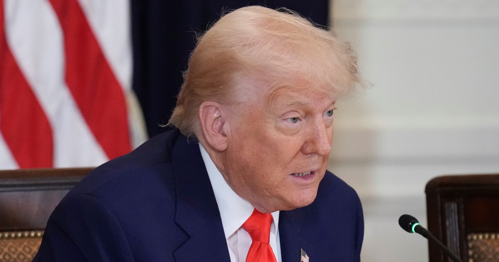 Donald Trump sitzt im Anzug mit roter Krawatte vor einem Mikrofon, im Hintergrund ist eine US-Flagge zu sehen.