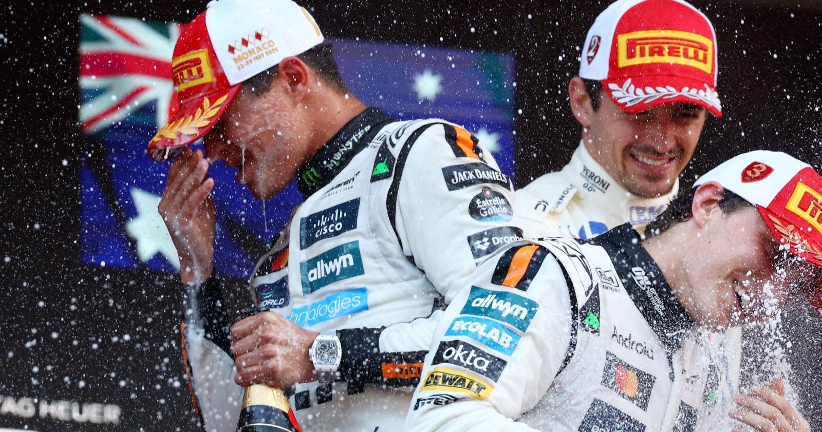 Lando Norris spritzt mit Champagner, neben ihm Oscar Piastri und Charles Leclerc auf dem Podium des Formel-1-Rennens in Monaco 2025, alle tragen Pirelli-Kappen und Rennanzüge.