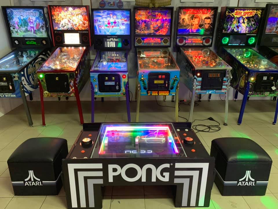 Ein Raum mit einer Sammlung klassischer Flipperautomaten an der Wand, darunter Titel wie "Alice Cooper's Nightmare Castle", "The Addams Family", "Star Wars" und "The Machine: Bride of Pin·Bot". Im Vordergrund steht ein moderner, beleuchteter Atari PONG-Tisch mit zwei schwarzen Hockern, ebenfalls mit Atari-Logo. Der Raum ist gut beleuchtet und vermittelt den Eindruck eines Retro-Spielhallenambientes.