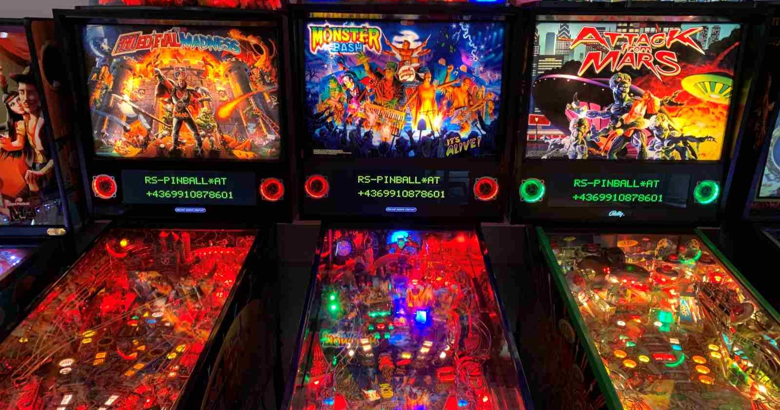 Drei beleuchtete Flipperautomaten nebeneinander in einer Spielhalle. Die Titel der Maschinen sind von links nach rechts: "Medieval Madness", "Monster Bash" und "Attack from Mars". Die Spielflächen sind bunt beleuchtet und zeigen jeweils thematisch passende Designs und Figuren.