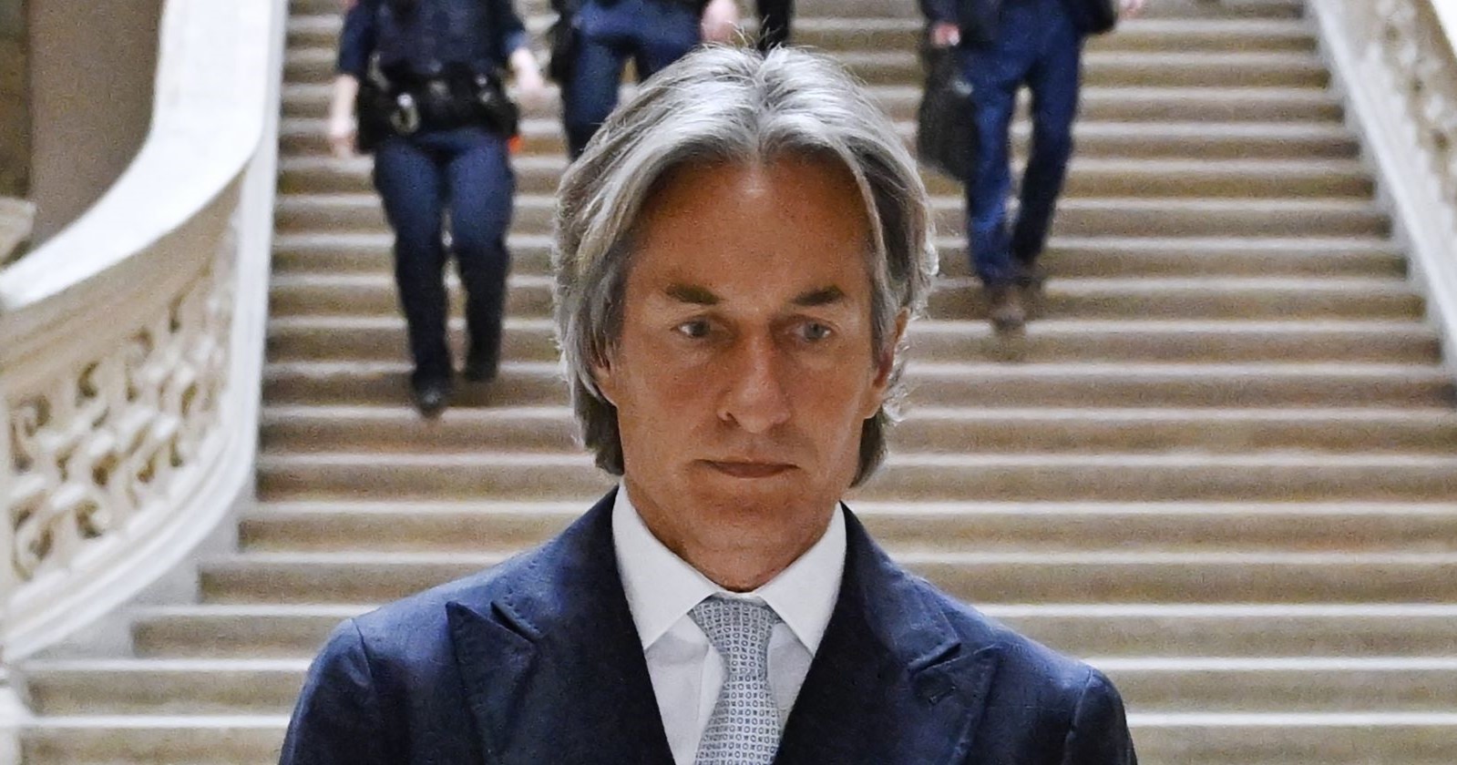 Karl-Heinz Grasser in Anzug und Krawatte geht eine monumentale Steintreppe hinunter, im Hintergrund mehrere uniformierte Personen – ernster Gesichtsausdruck.