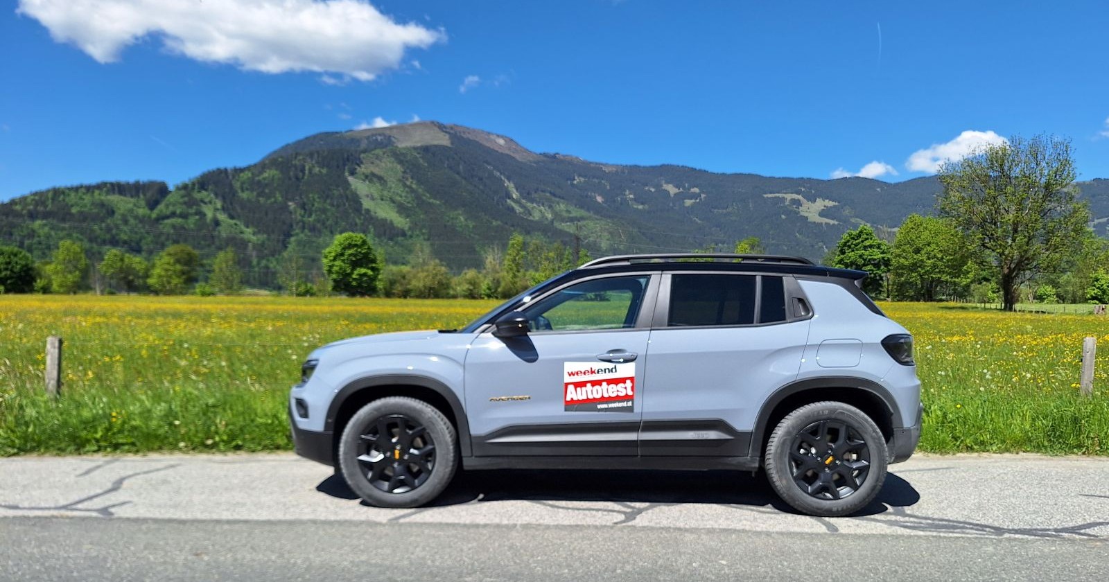Ein hellblauer Jeep Avenger parkt auf einer Straße. Im Hintergrund sind eine Wiese mit gelben Blumen zu sehen und einige Obstbäume. Dahinter sieht man auch - nicht sehr hohe - Berge - die bewaldet sind.