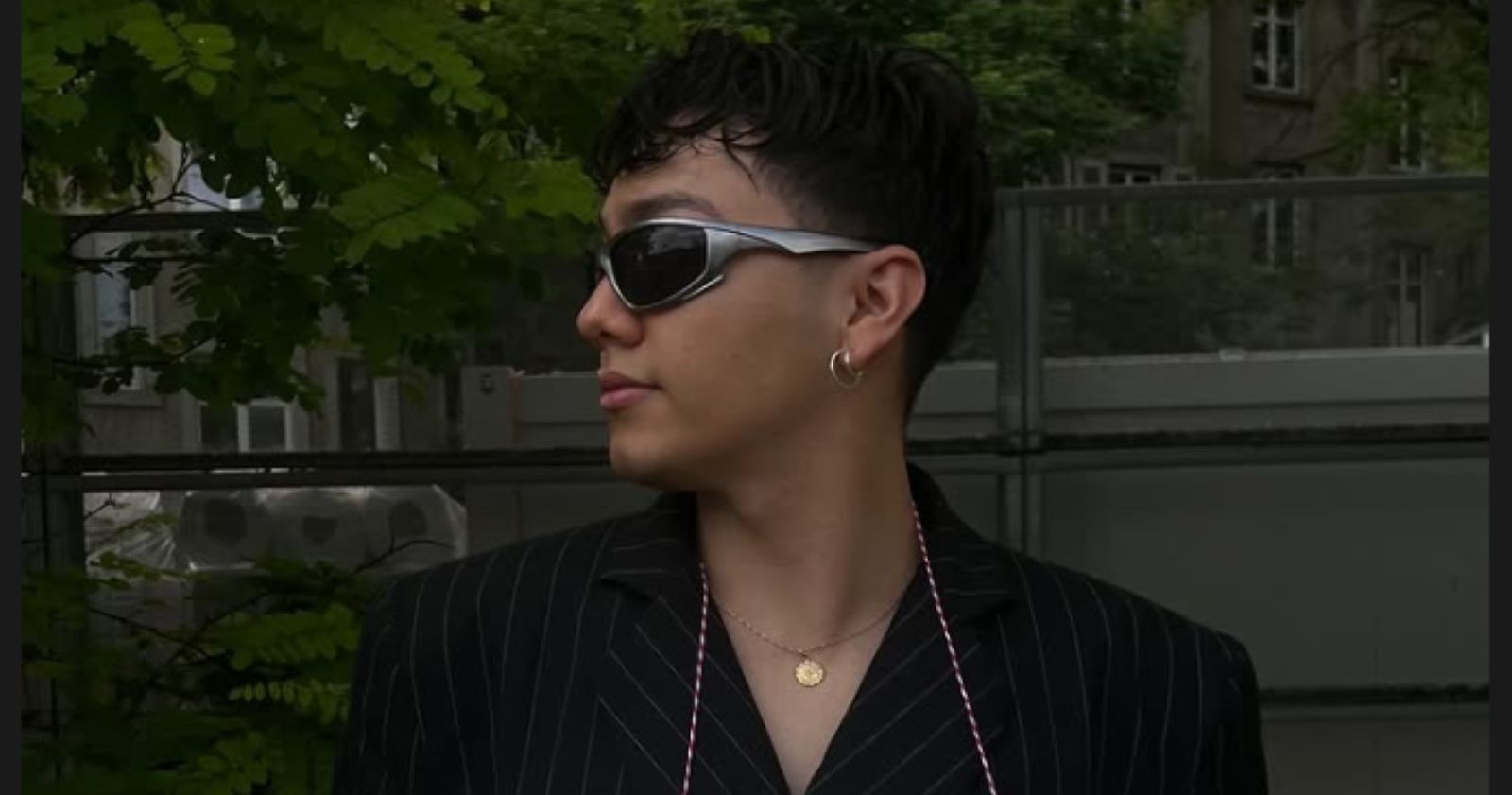 Johannes "JJ" Pietsch schaut zur Seite, trägt ein schwarzes Hemd, silberfarbene Ohrringe, eine goldfarbene Kette und eine Sonnenbrille