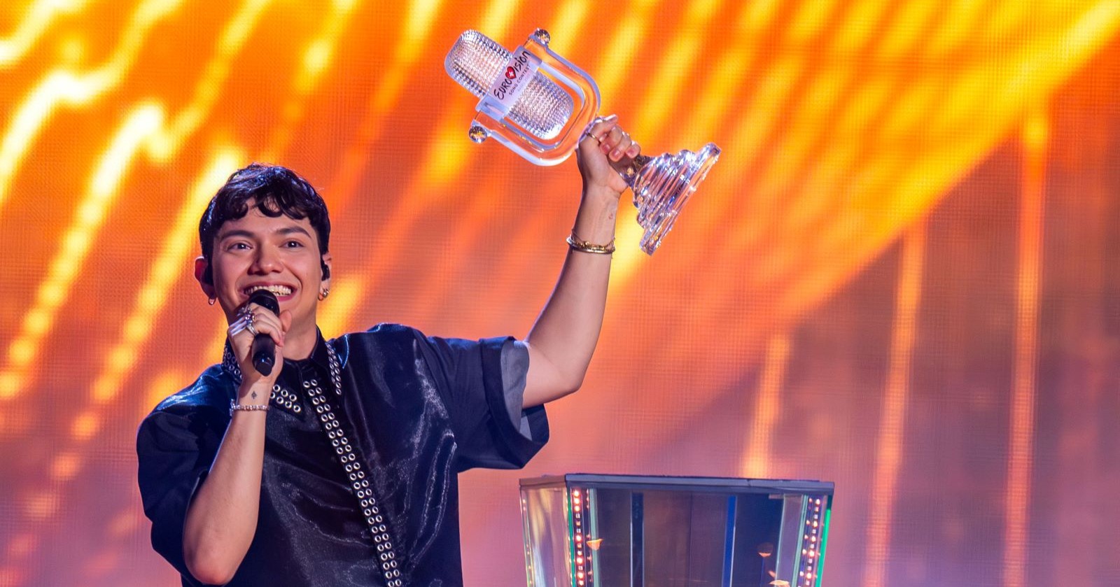 JJ, lächelnd mit Mikrofon in der linken Hand hält mit der rechten Hand die gläserne ESC-Trophäe in die Höhe. Im Hintergrund leuchten orange-gelbe Lichter auf der Bühne.