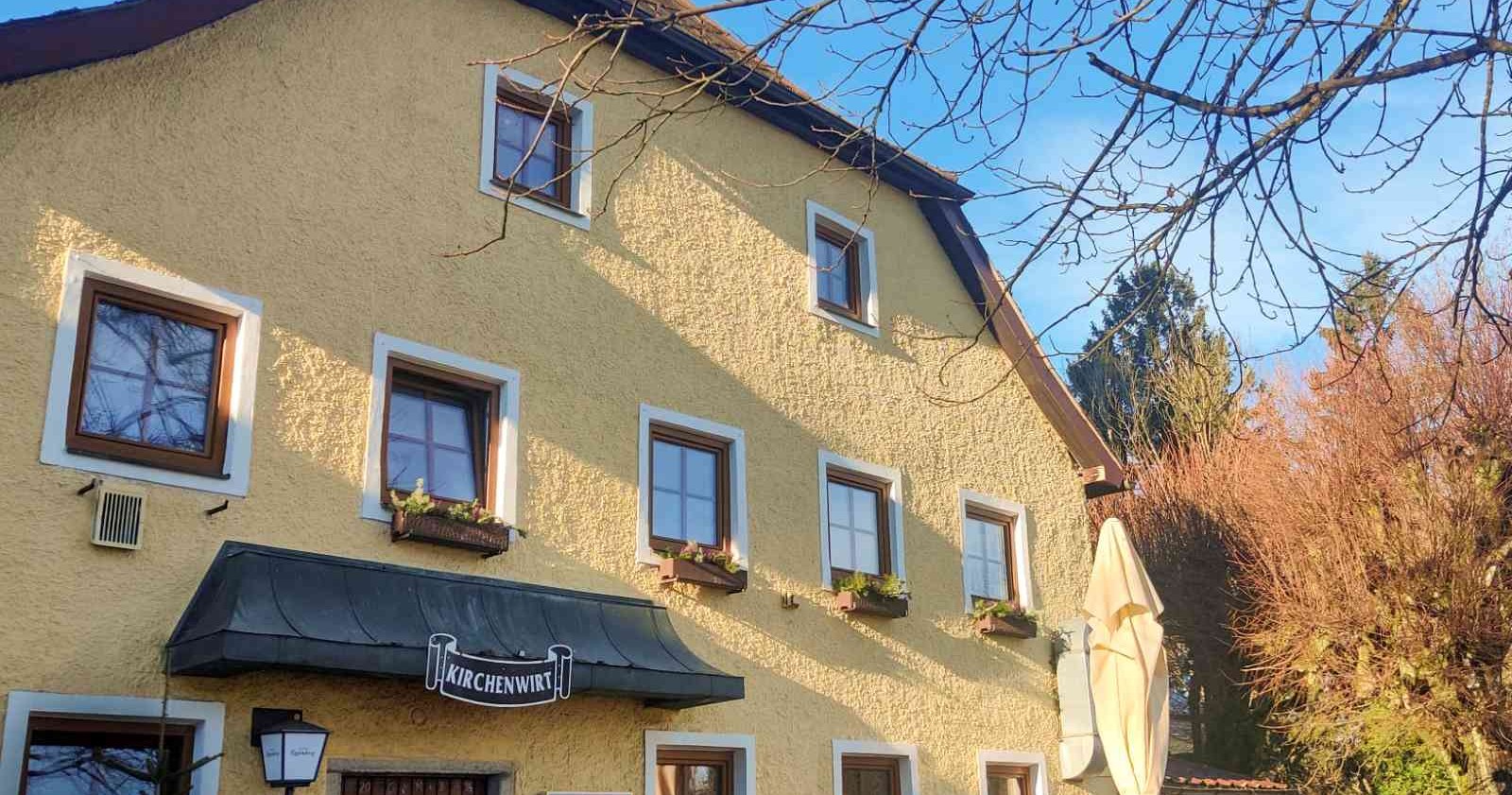 Gelbes, traditionelles Gasthaus mit dem Namen „Kirchenwirt“ auf einem Schild über dem Eingang. Das Gebäude hat mehrere Fenster mit Blumenkästen und ist von kahlen Bäumen sowie herbstlich gefärbtem Gehölz umgeben. Der Himmel ist klar und blau.