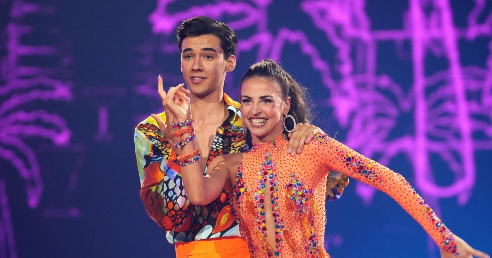 Diego Pooth und seine Tanzpartnerin Ekaterina Leonova lächeln auf der "Let's Dance"-Bühne im Finale 2025. Beide tragen bunte, glitzernde Kostüme, die Kulisse ist violett beleuchtet.