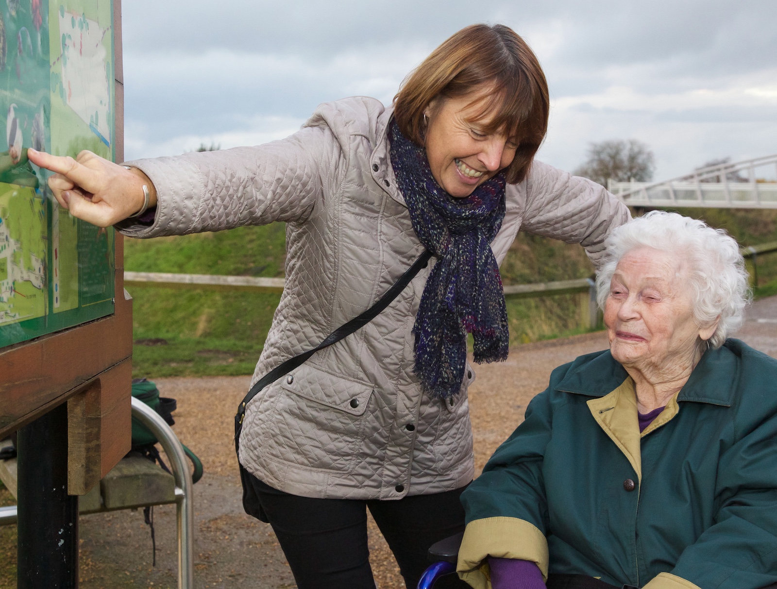 Ein Frau geht mit einer Seniorin spazieren und zeigt ihr etwas an einer Tafel.