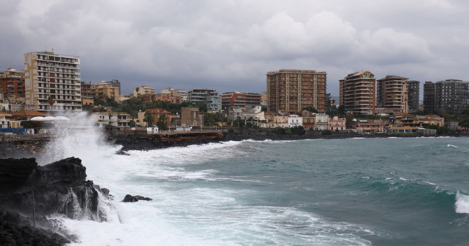 Küste der sizilianischen Stadt Catania bei Sturm: hohe Wellen schlagen gegen dunkles Lavagestein, im Hintergrund mehrstöckige Wohnhäuser unter grauem Himmel.