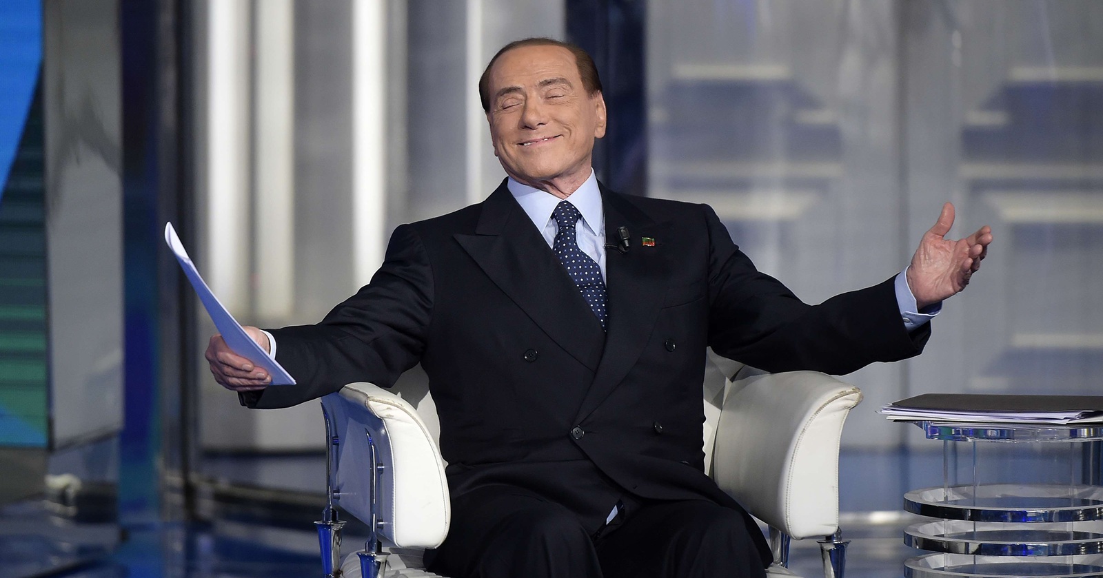 Silvio Berlusconi sitzt im TV-Studio und breitet gönnerhaft die Arme aus