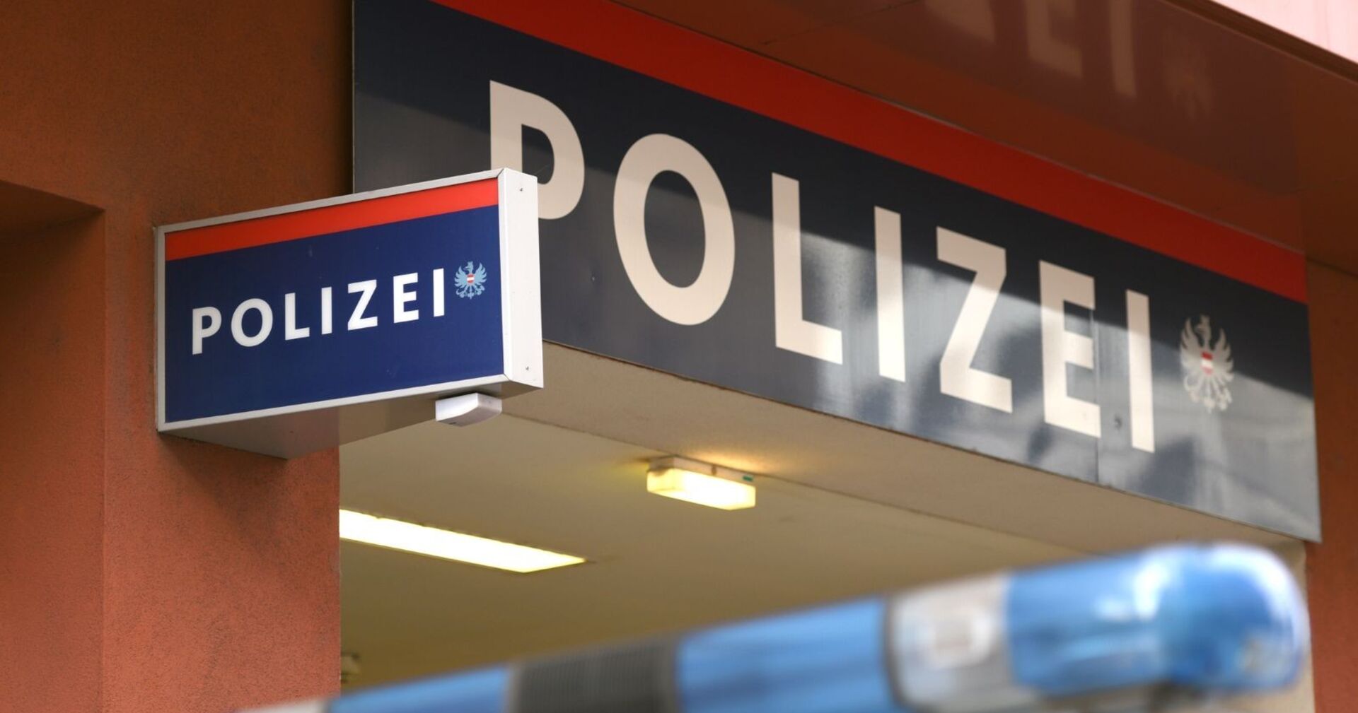 Eingangsbereich einer Polizeiinspektion in Österreich mit blau-rotem Polizeischriftzug und Blaulicht eines Polizeiwagens im Vordergrund.