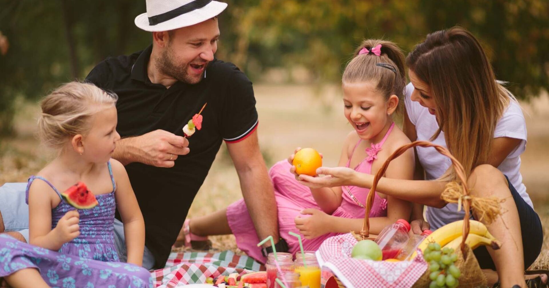 Eine fröhliche Familie beim Picknick im Freien. Zwei Kinder und ihre Eltern sitzen auf einer Decke, essen frisches Obst und lachen miteinander. Der Vater trägt einen weißen Hut und hält einen Obstspieß, während die Mutter und eine Tochter eine Orange betrachten. Im Vordergrund steht ein Picknickkorb mit Bananen, Trauben und Getränken. Die Szene findet an einem sonnigen Tag im Grünen statt.