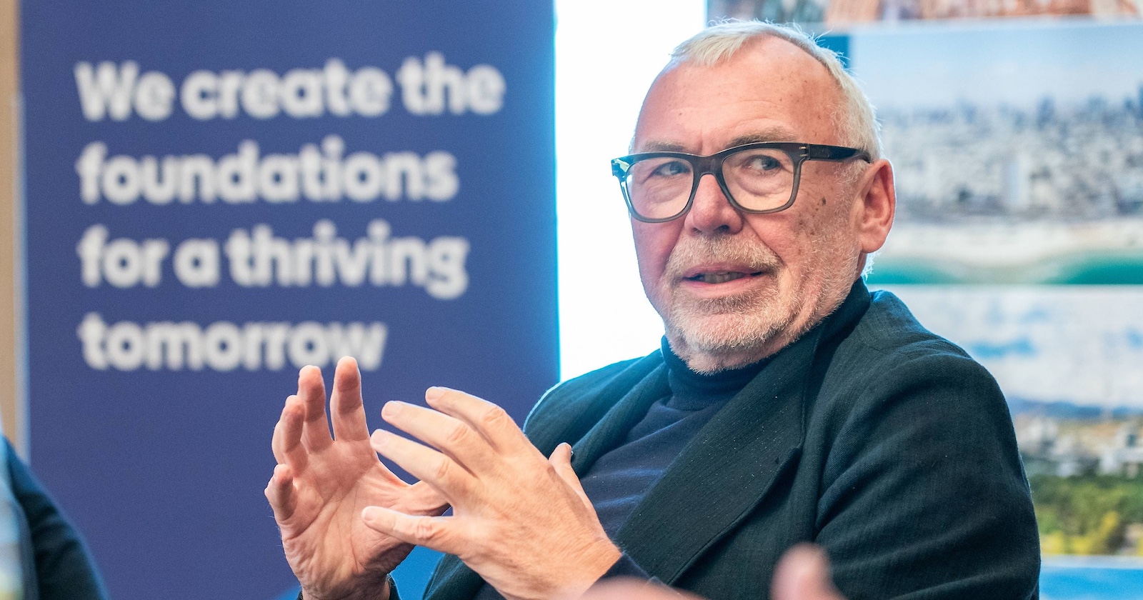 Alfred Gusenbauer spricht gestikulierend bei einer Veranstaltung, im Hintergrund ein Poster mit dem Schriftzug „We create the foundations for a thriving tomorrow“.