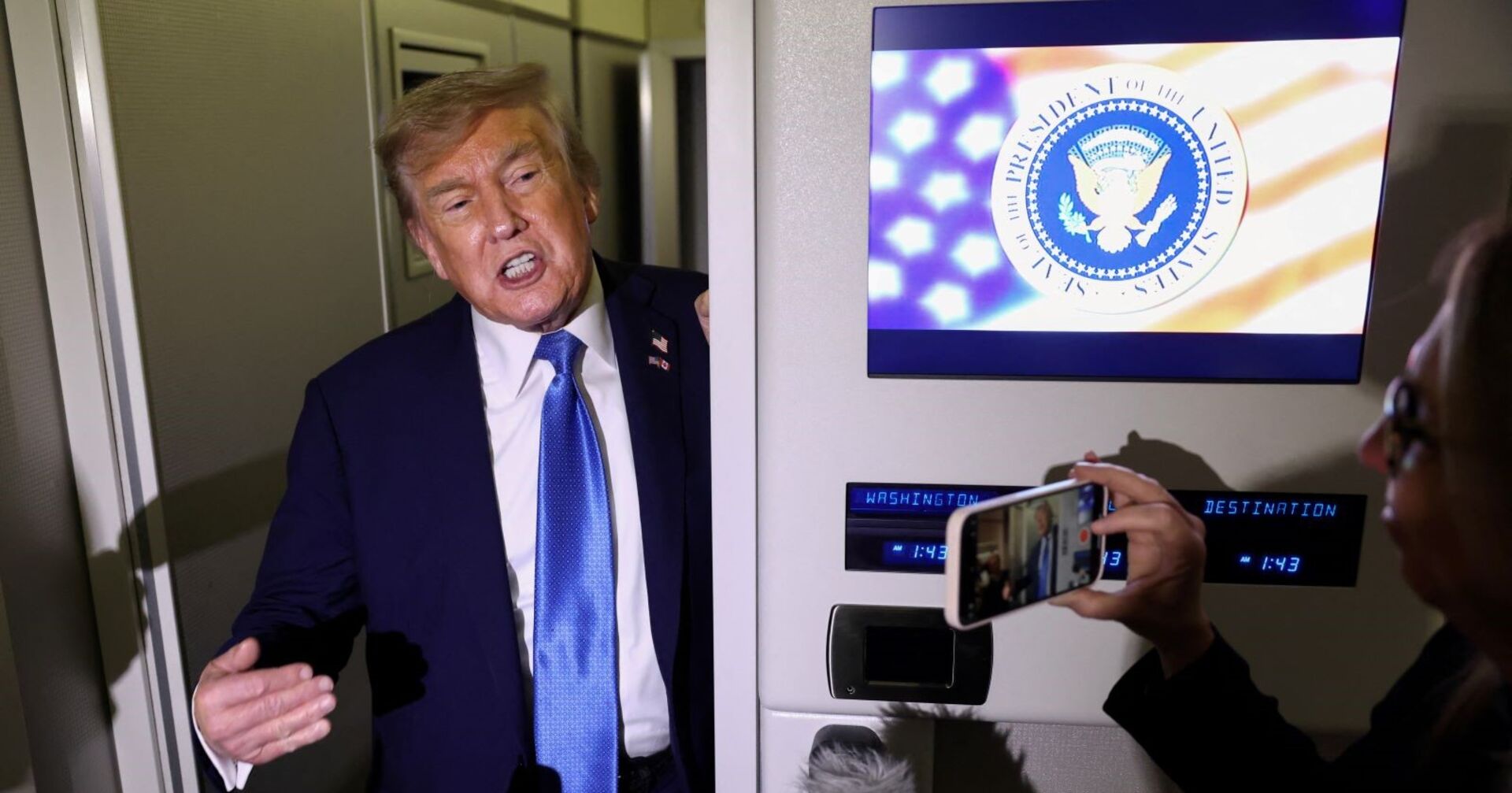 Donald Trump spricht in einem Flugzeug mit Medienvertretern, neben ihm ein Bildschirm mit dem Präsidentensiegel der USA. Eine Person filmt ihn mit dem Smartphone.