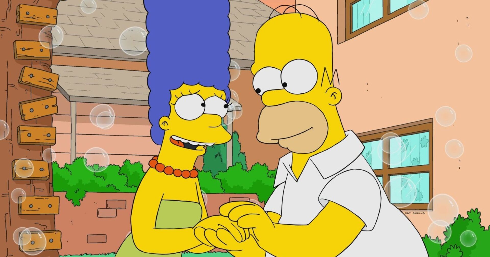 Marge und Homer Simpson halten sich liebevoll die Hände und blicken sich in die Augen, mit Seifenblasen im Vordergrund und ihrem Haus im Hintergrund.